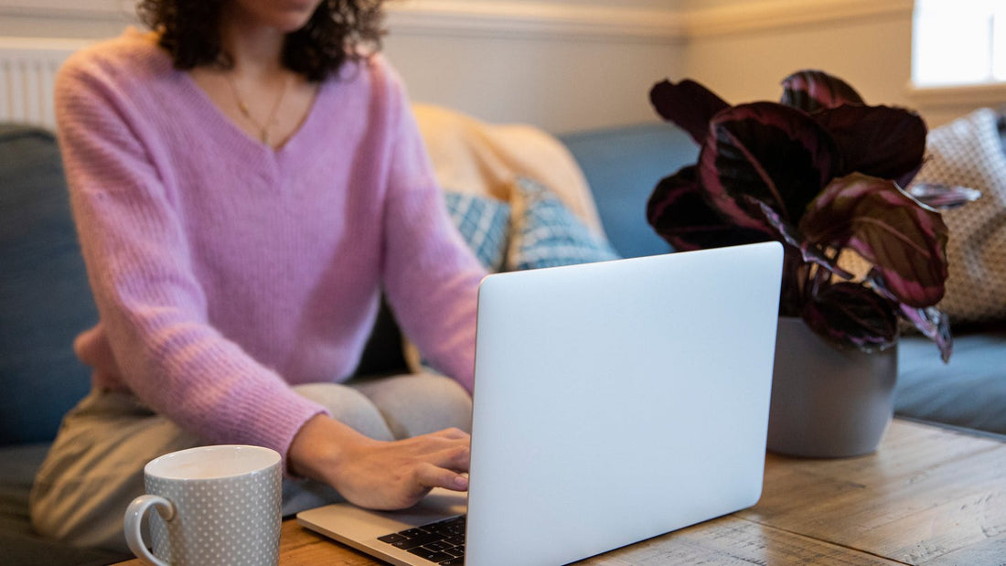 A person in a soft sweater working on a laptop from a home couch, with a coffee mug and a houseplant on the wooden table