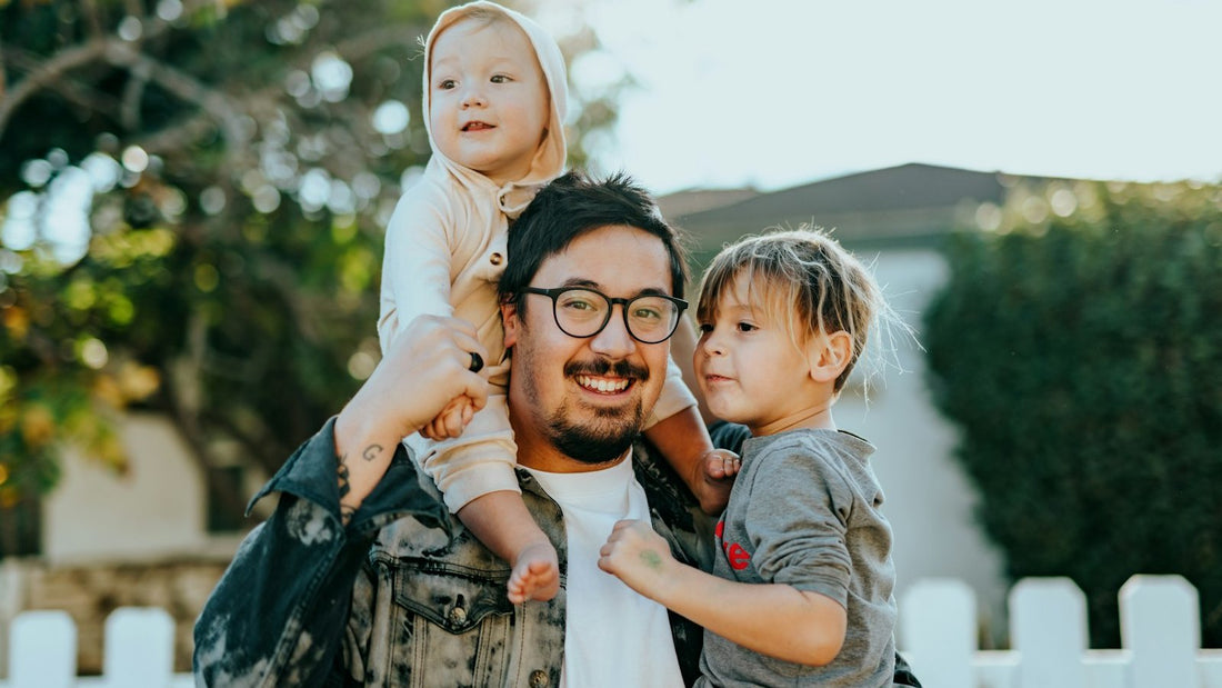 A smiling young father outdoors holding a toddler on his shoulders and a small child in his arm, in front of a white picket fence.