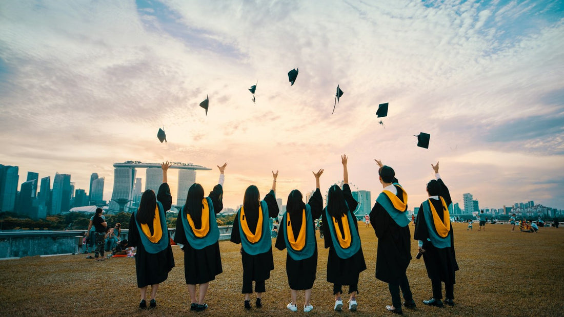 Six university graduates in caps and gowns stand on a field with arms raised, tossing their mortarboards into a warm sunset sky over a city skyline