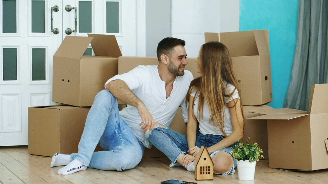 Smiling couple sitting on the floor of a new home, surrounded by cardboard moving boxes, with a small wooden house and a green plant beside them