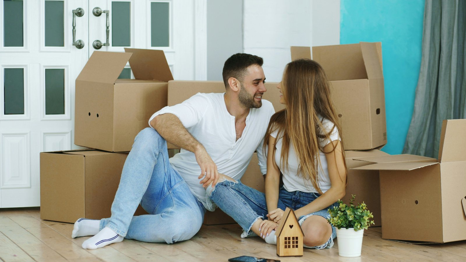 Smiling couple sitting on the floor of a new home, surrounded by cardboard moving boxes, with a small wooden house and a green plant beside them