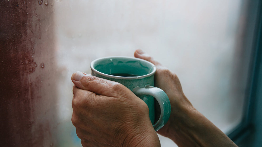 A pair of hands holds a warm green ceramic mug against a rainy window — a quiet moment of self-care.