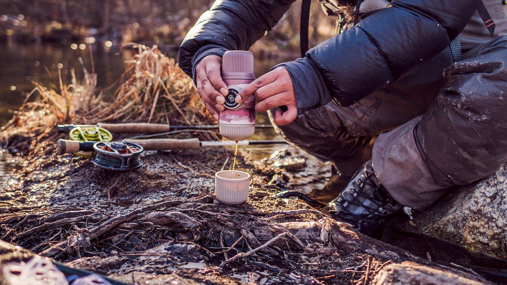 Hands pulling a hand-pump portable espresso shot into a small cup on a riverbank at golden hour, with a fly fishing rod and net resting on the ground beside the brewer