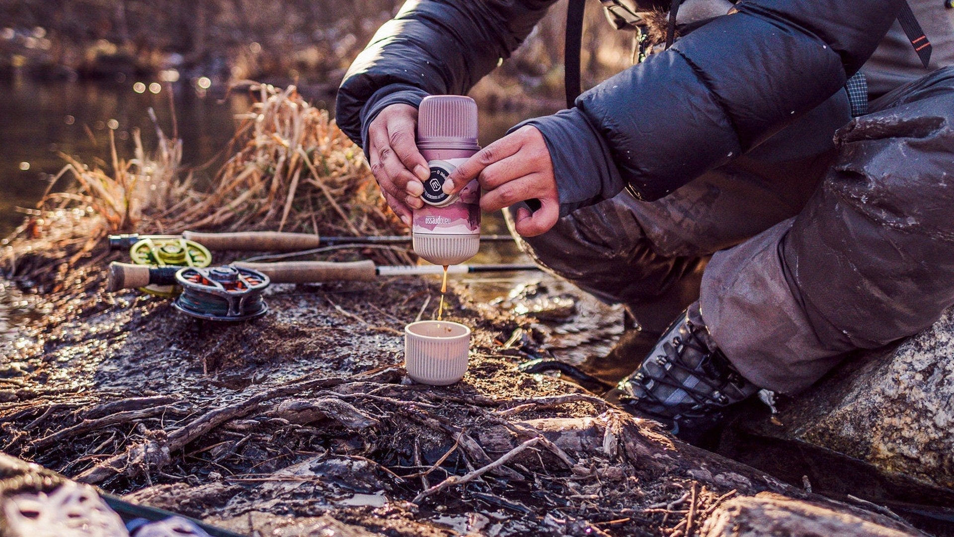 Hands pulling a hand-pump portable espresso shot into a small cup on a riverbank at golden hour, with a fly fishing rod and net resting on the ground beside the brewer