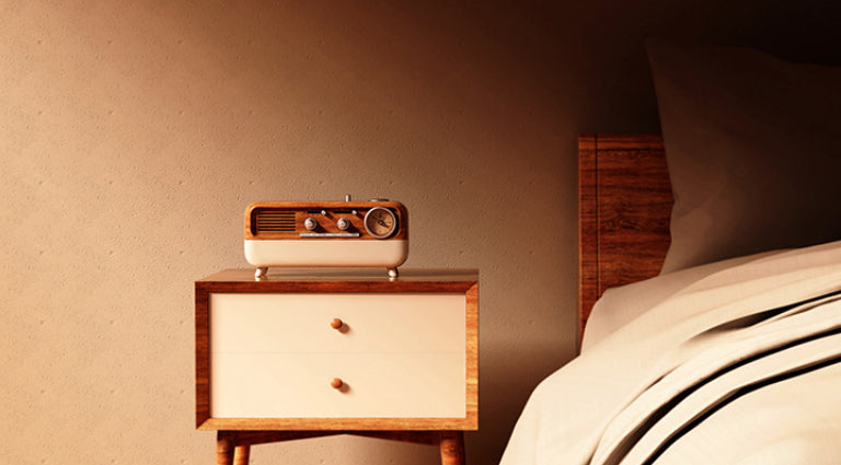 Retro walnut-grain Bluetooth speaker sitting on a wooden nightstand next to a bed in a warm bedroom