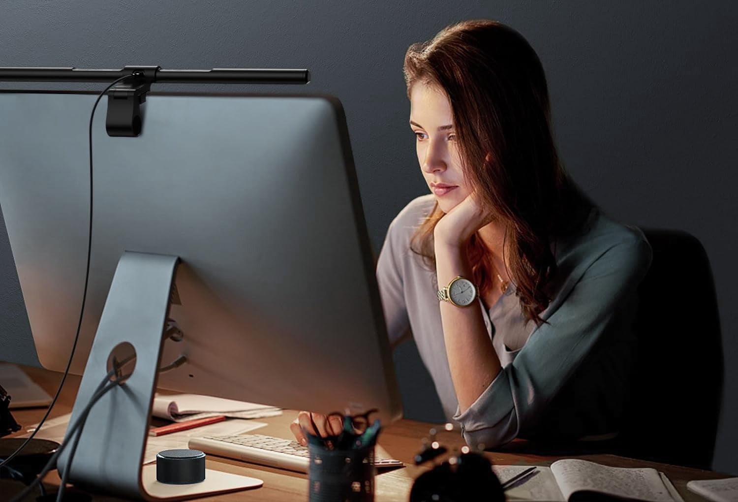 Halo Monitor Light Bar mounted on top of a desktop monitor in a dimly lit home office, with a young woman working at the desk and the round wireless remote dial visible on the desk surface