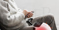 Older man in cream sweater scrolling his phone in an armchair while a Smart Knee Massager with red-light therapy warms his knee.