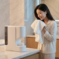 Hot Towel Oshibori Dispenser on a marble bathroom counter — a woman in cream pajamas presses a freshly steamed cotton towel to her face, smiling, the dispenser beside her on the vanity.