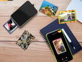 Portable instant photo printer on a wooden desk surrounded by scattered freshly printed photos, a notebook with a smartphone showing the printing app, a pen and a small wooden pile of pebbles in a flatlay composition