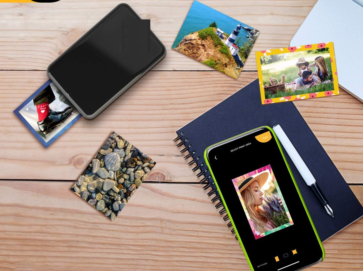 Portable instant photo printer on a wooden desk surrounded by scattered freshly printed photos, a notebook with a smartphone showing the printing app, a pen and a small wooden pile of pebbles in a flatlay composition