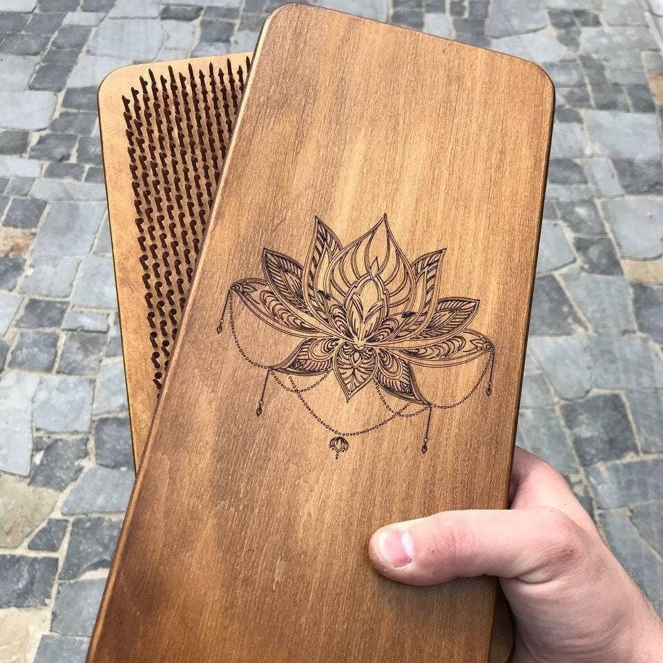 Handcrafted wooden Sadhu nail board with engraved lotus mandala held in a hand outdoors on a cobblestone street, with a second board behind it showing the metal nails on its acupressure surface