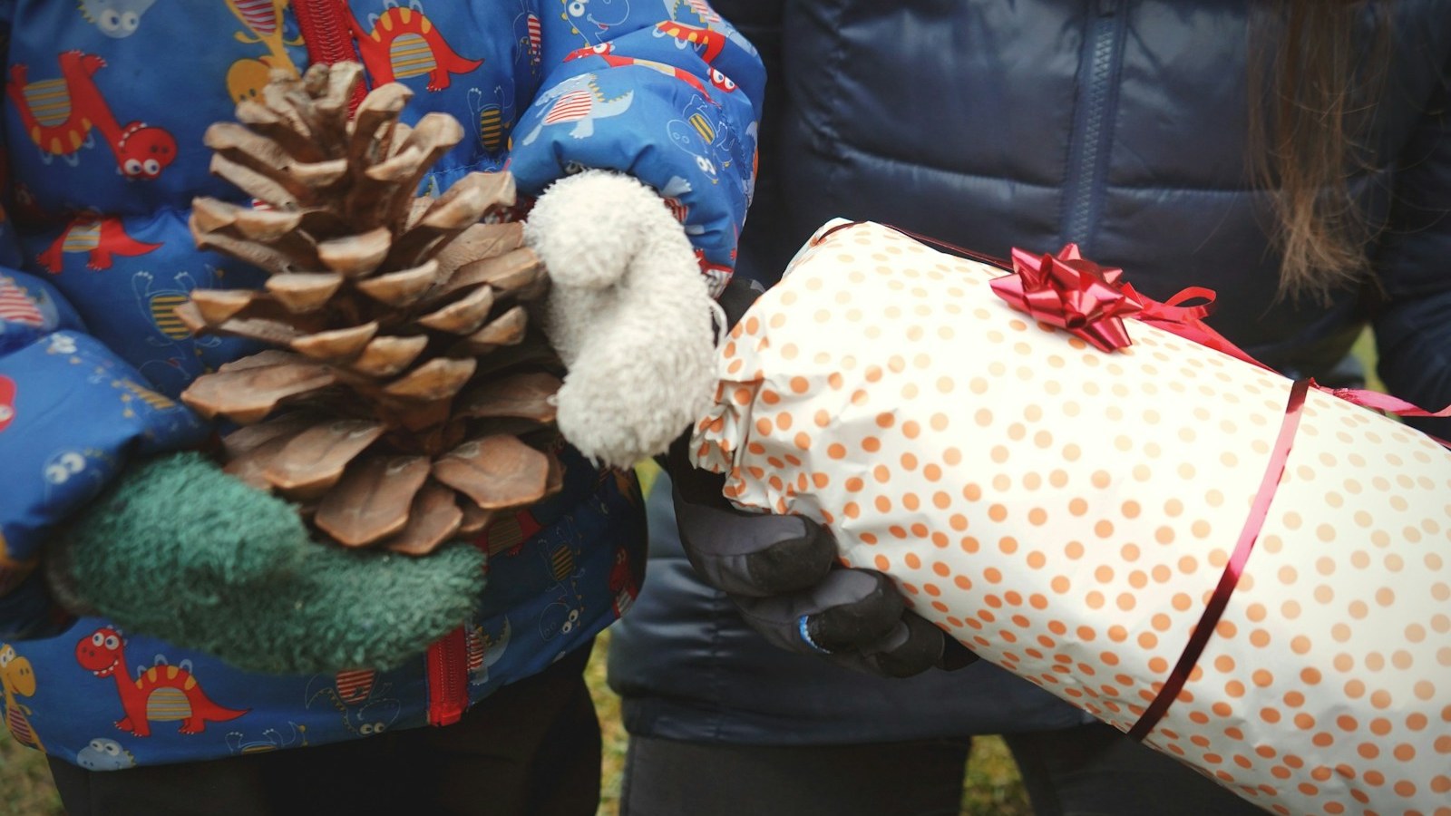 Outdoor scene of a small child in a blue dinosaur-print winter coat and mittens holding a large pine cone, exchanging with an adult in a dark navy coat holding a polka-dot wrapped gift with a red bow